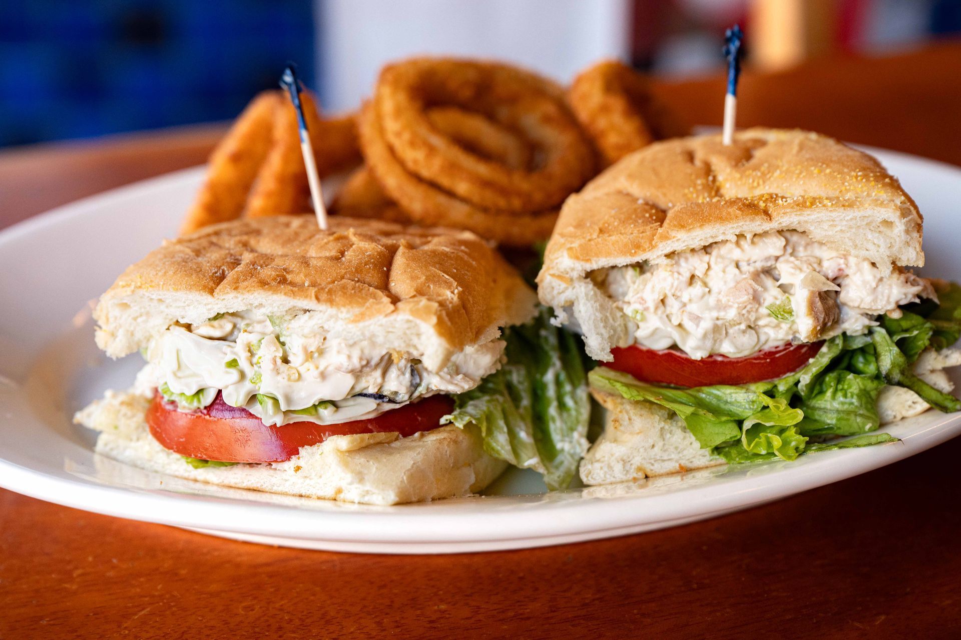 Two sandwiches with lettuce and tomato on a plate, served with onion rings in the background