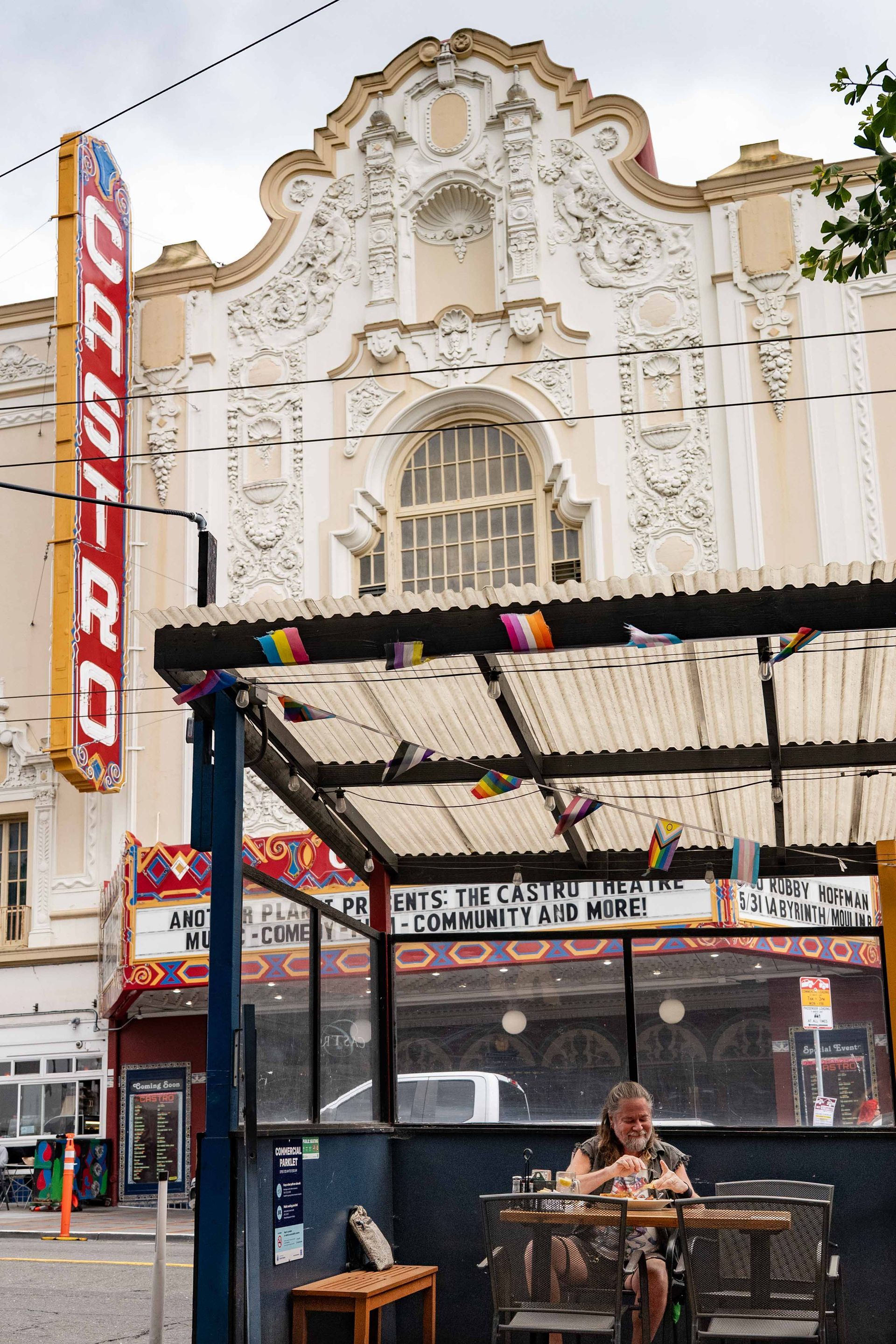 Outdoor café under an ornate white theater façade with colorful signage and a seated person at a table.