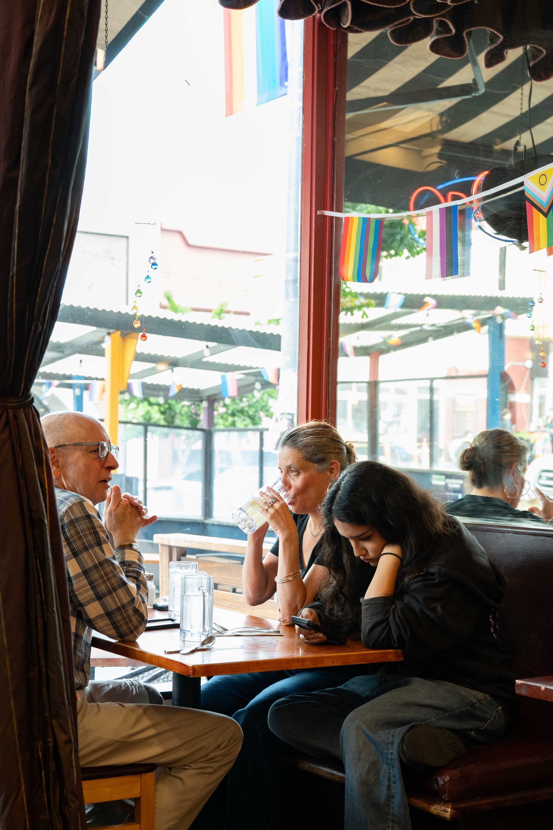People seated at a table in a café by a window, with rainbow flags hanging above.