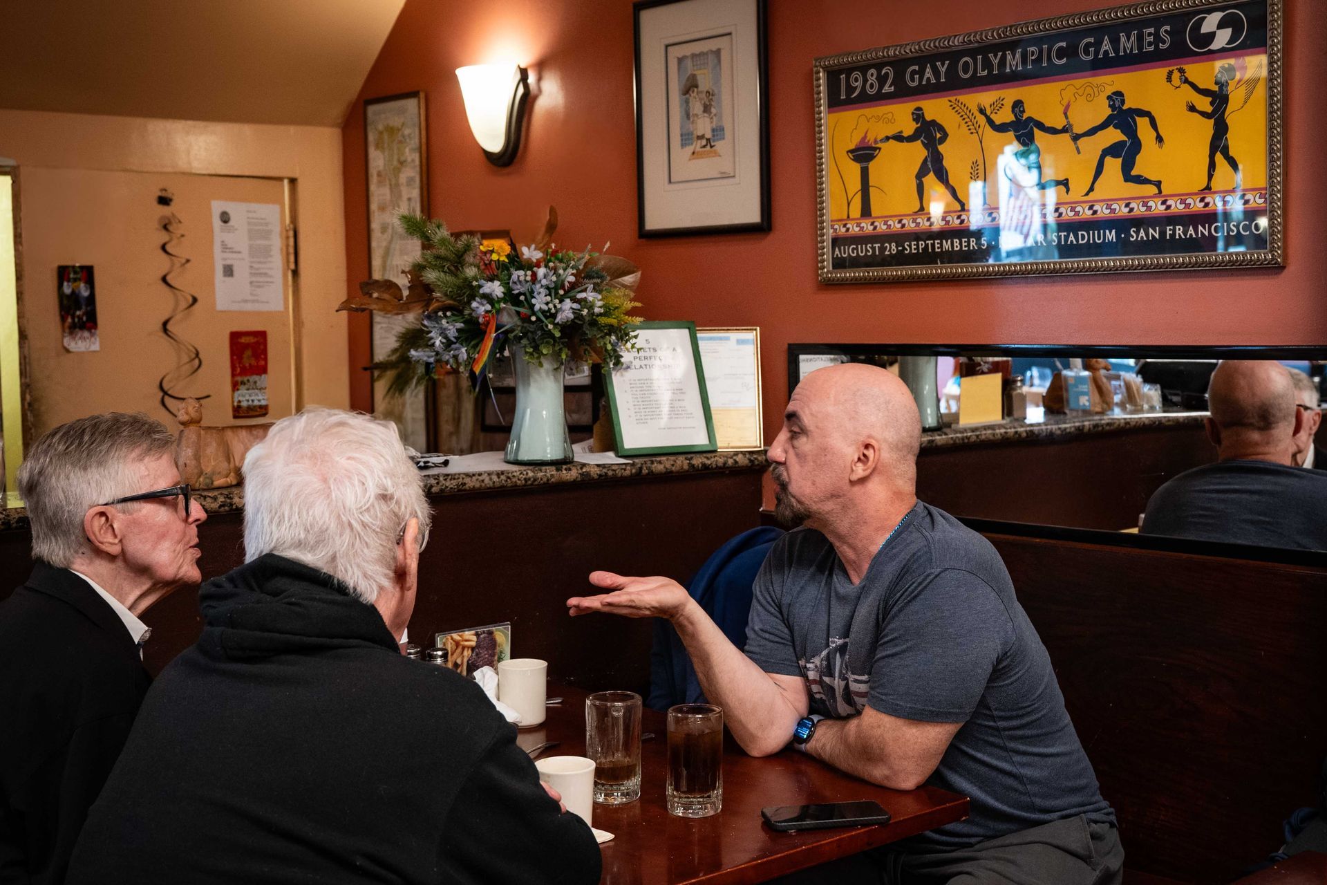 Three people talk at a pub table beneath sports wall art and a mirror