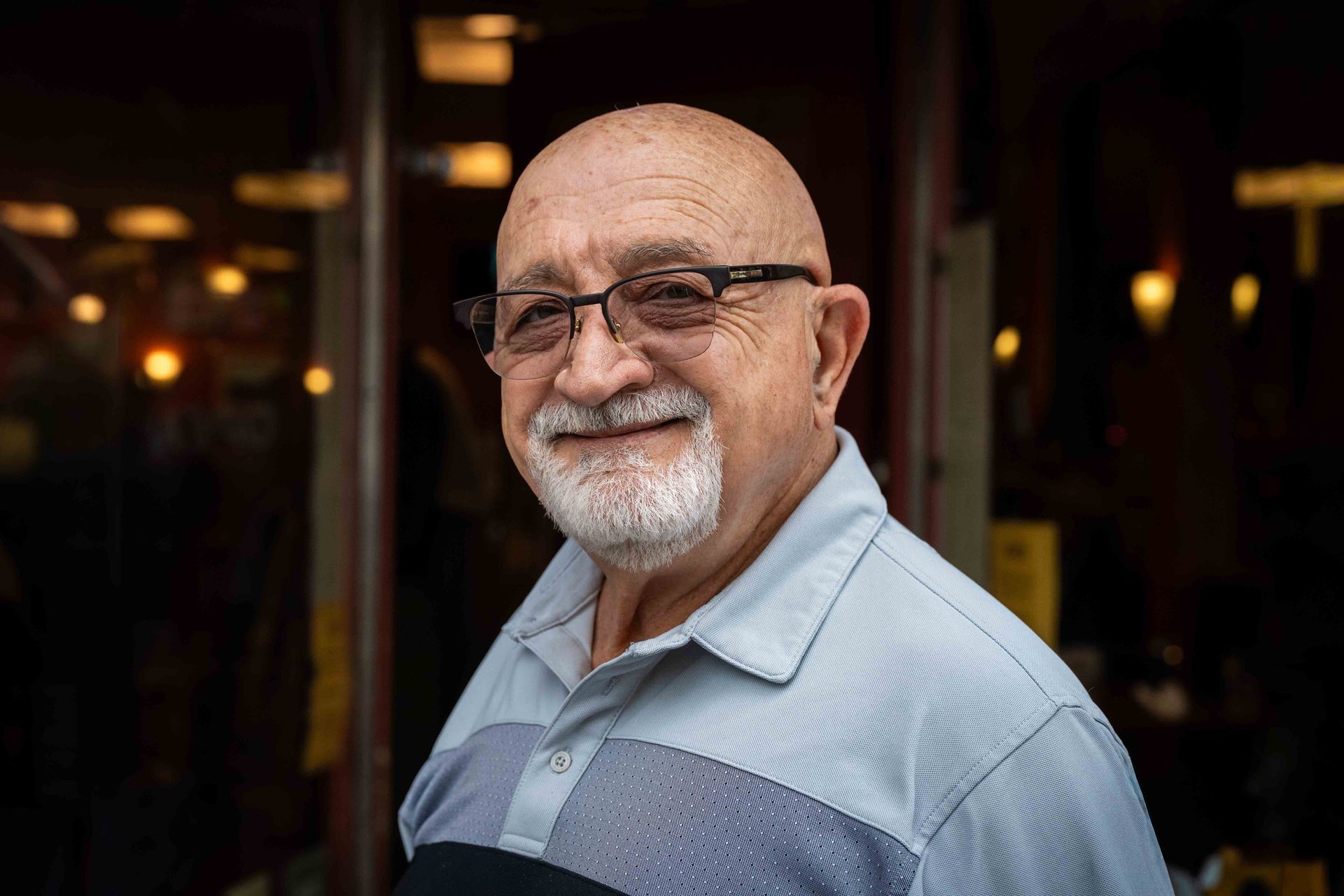 Smiling bald man with glasses in a light blue shirt standing outdoors at dusk