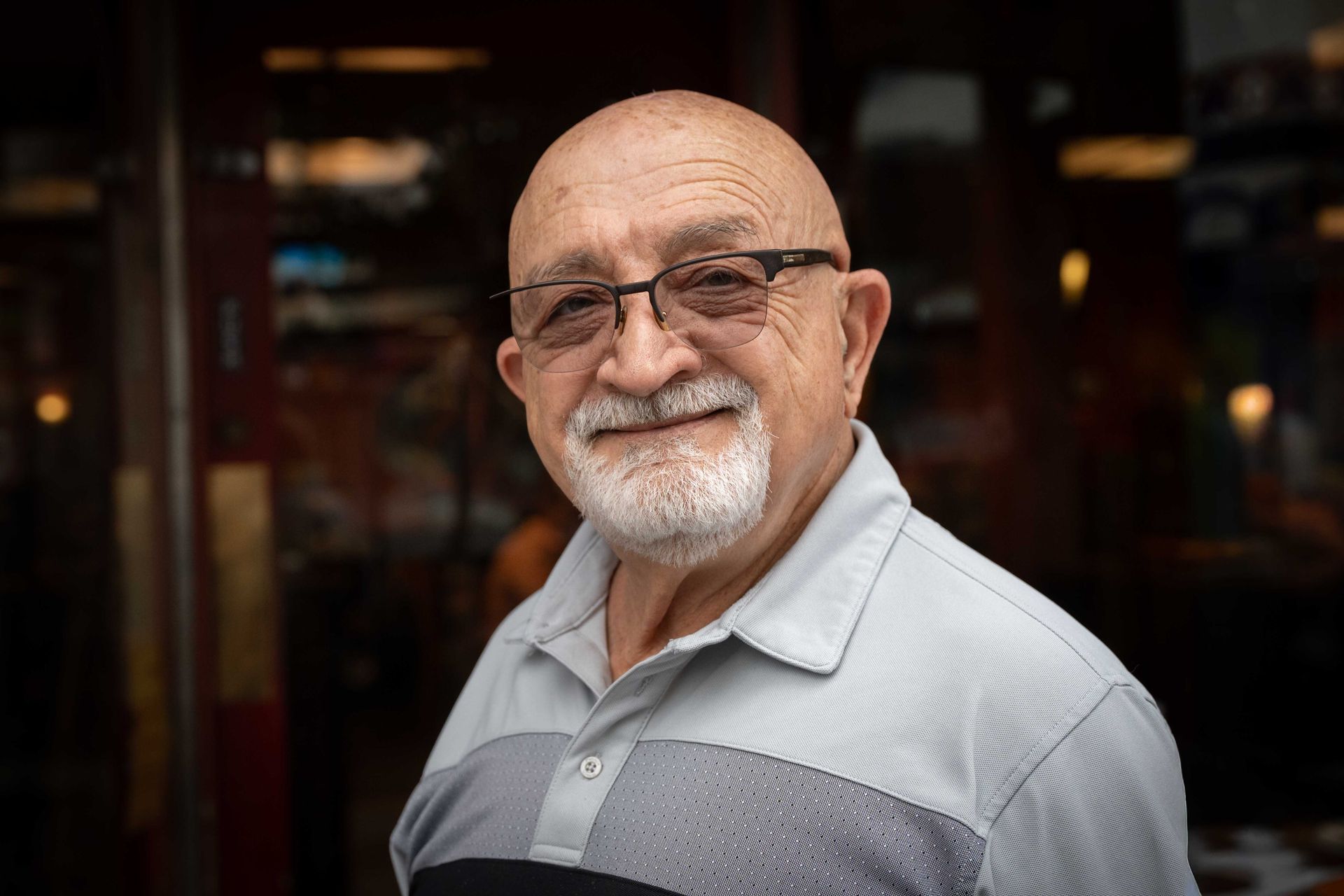 Smiling bald man with glasses and white beard in a light gray shirt, standing indoors with blurred lights behind him