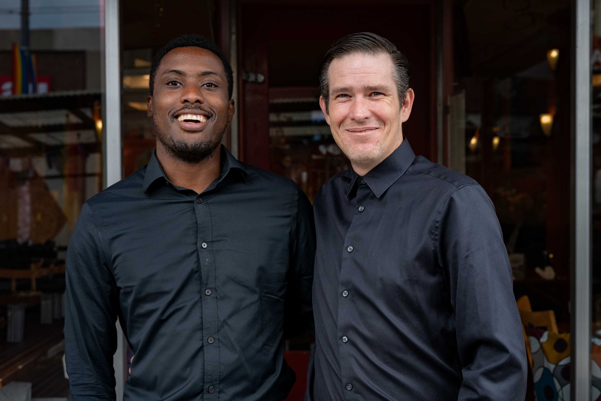Two men standing together indoors, both wearing dark shirts and smiling at the camera.