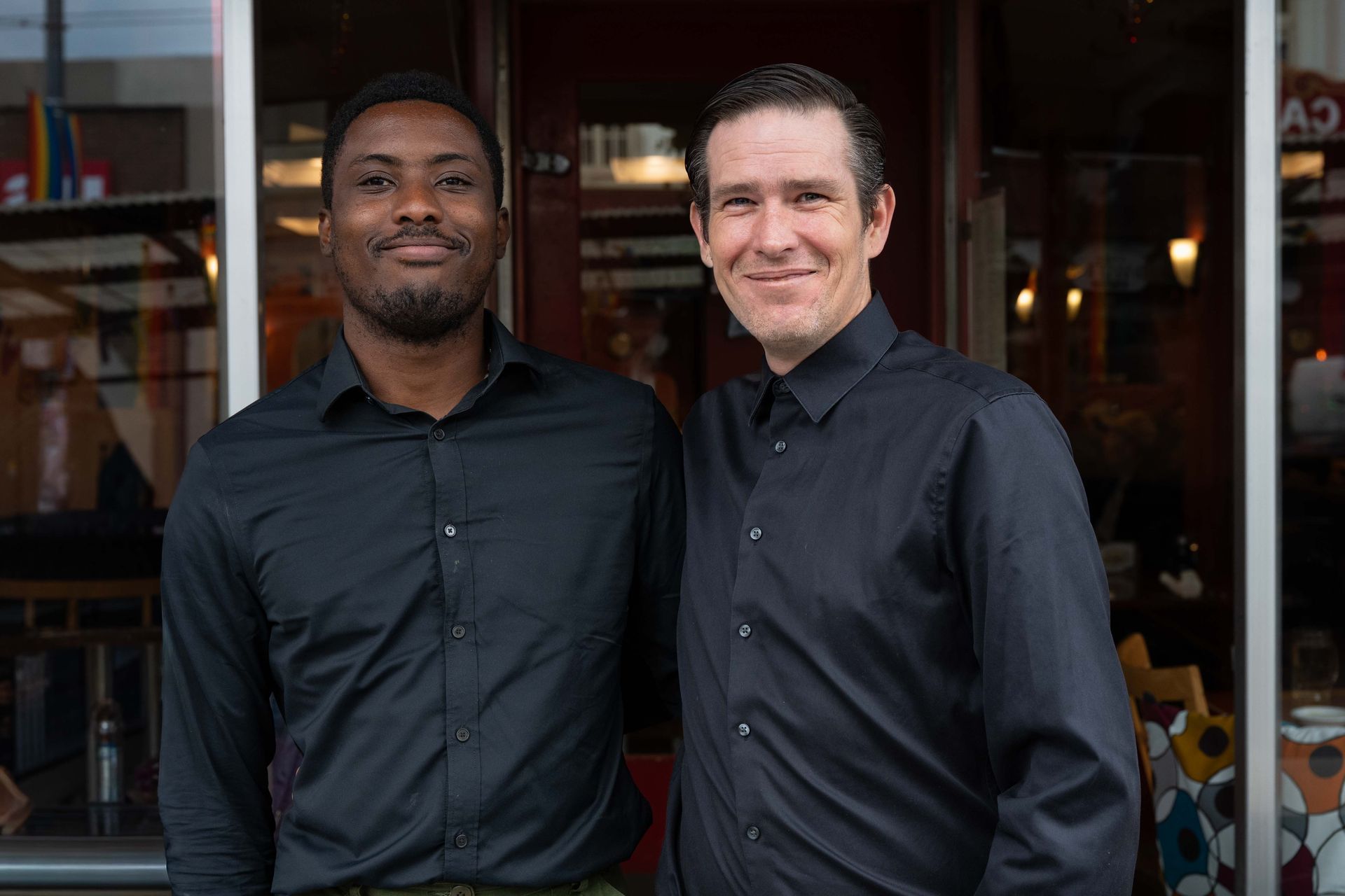 Two men in dark shirts standing side by side in front of a storefront, smiling at the camera.