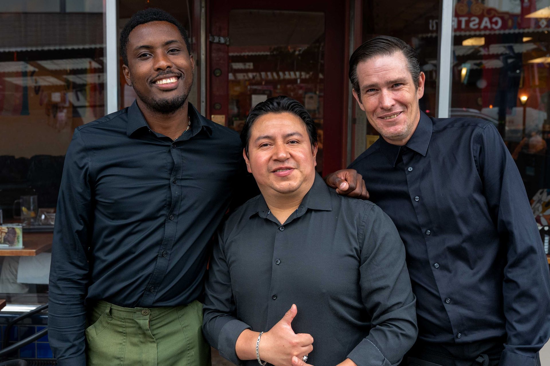 Three men in dark shirts smiling and standing together outside a storefront.