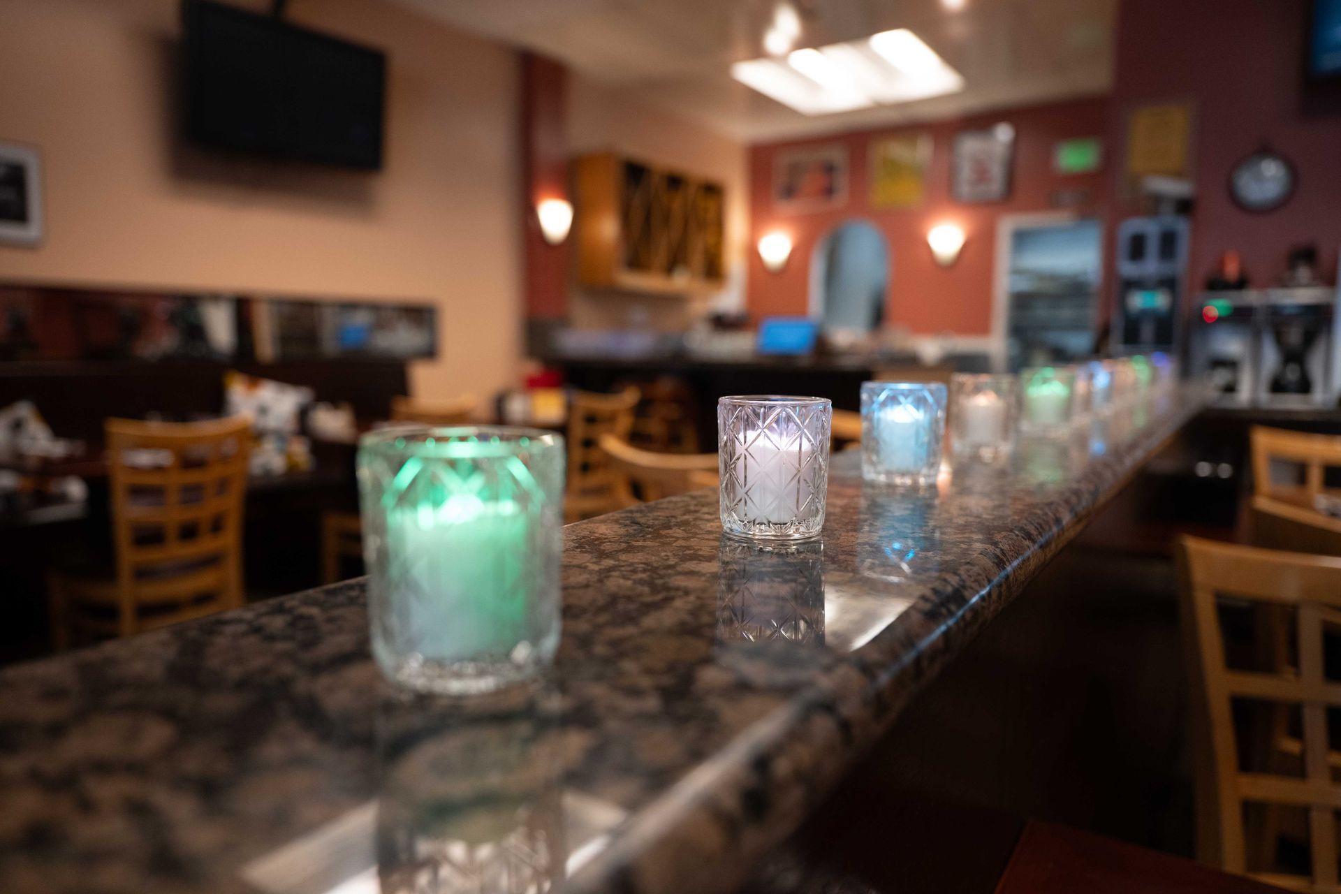 Colorful candles in glass holders line a polished bar in a dimly lit restaurant.