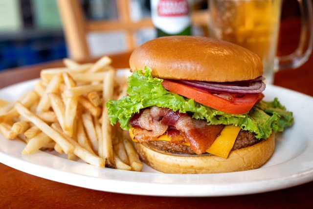 Burger with lettuce, tomato, bacon, and cheese on a plate with French fries and drinks in the background