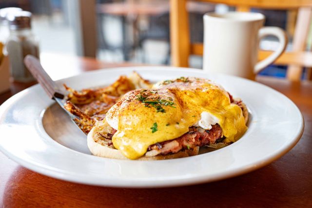 Cheesy eggs Benedict on a white plate with a fork beside a coffee cup on a restaurant table