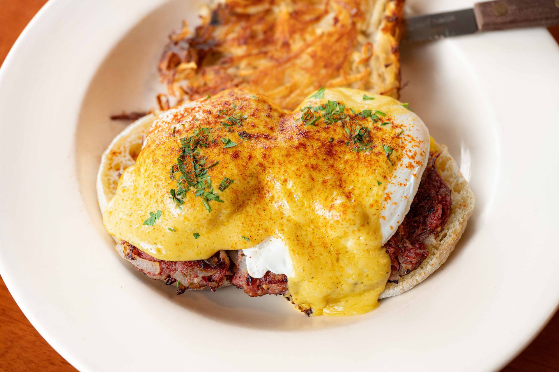 Eggs Benedict with hollandaise sauce and herbs on a white plate, with hash browns in the background
