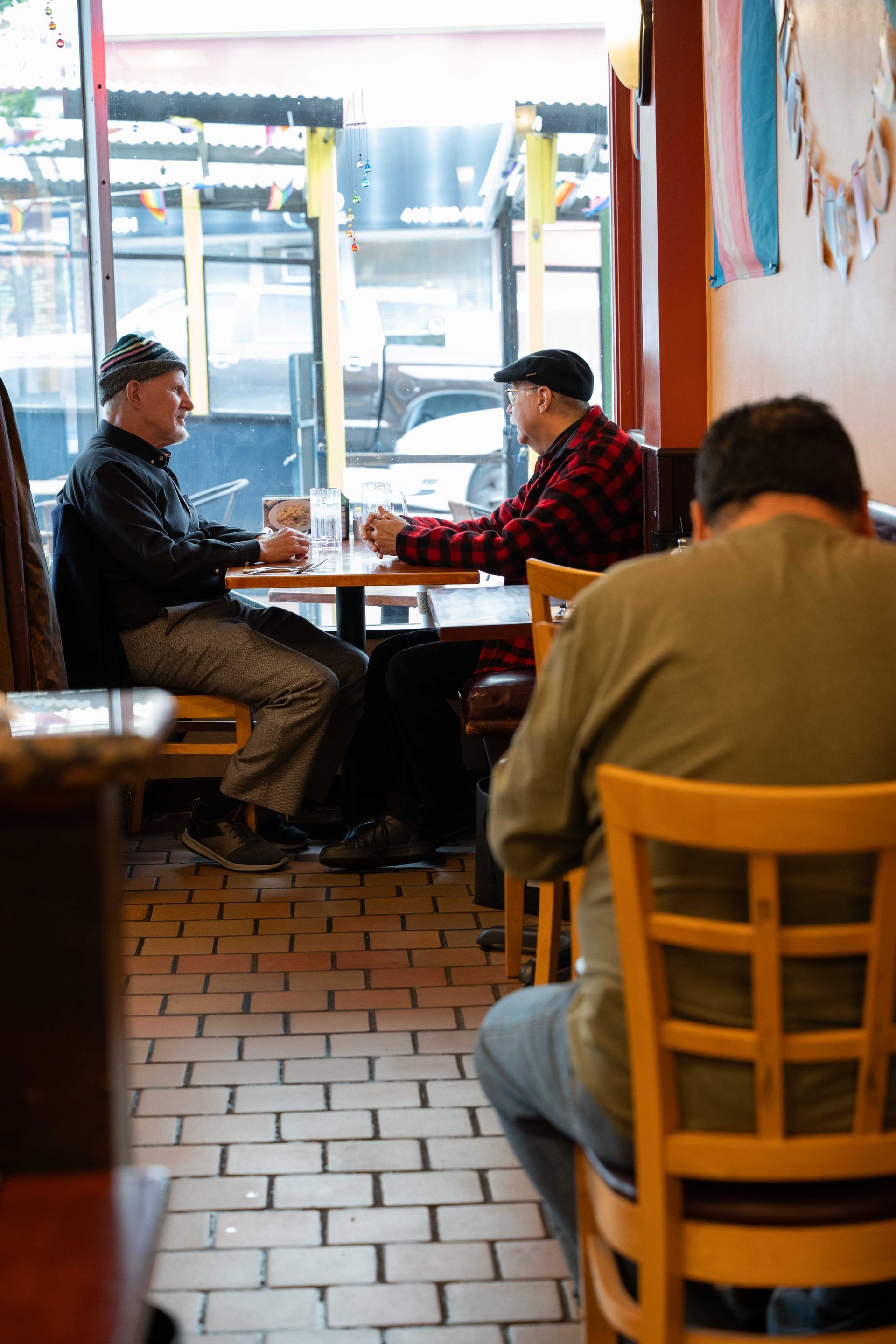 Two people sit at a café table near a window, with a person in a tan jacket seated in the foreground.