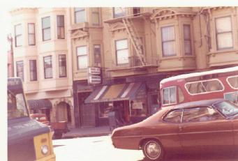 Street view of brownstone buildings and storefronts with parked cars in the foreground