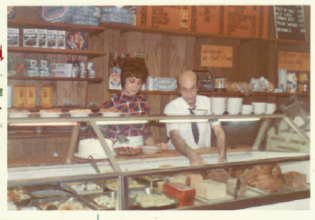 Two people behind a bakery counter with pastries and shelves of baked goods in the background