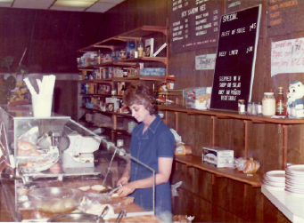 Woman in a diner kitchen serving food behind the counter, with menu boards and stacked plates.