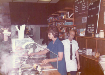 Worker behind a deli counter serving food while a man stands beside a chalkboard menu.