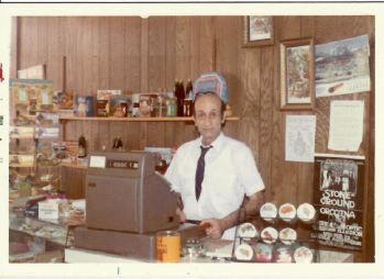 Shop clerk behind a counter in a wood-paneled store, wearing a white shirt and tie with displays of snacks and goods.