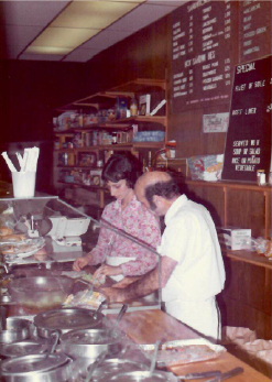 Two people preparing food behind a deli counter in a dimly lit restaurant.