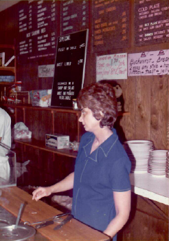 Woman behind a deli counter serving food, with menu boards and stacked plates in the background