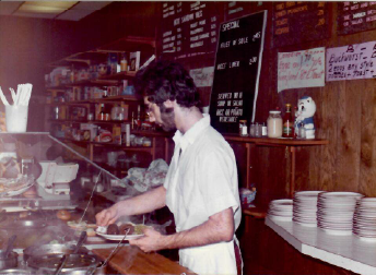Person in a white shirt serving food behind a diner counter with stacked plates and menu boards.