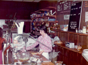 Woman behind glass bakery counter with pastries, chalkboard menu, and shelves in a small café