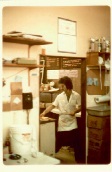 Person in white uniform working in a small lab or kitchen-like room with shelves and equipment.