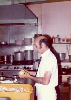 Chef preparing food in a commercial kitchen, holding dough near trays of baked goods.