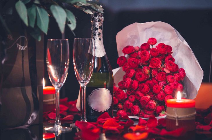 Uma garrafa de champanhe, duas taças, um buquê de rosas vermelhas e velas sobre uma mesa.