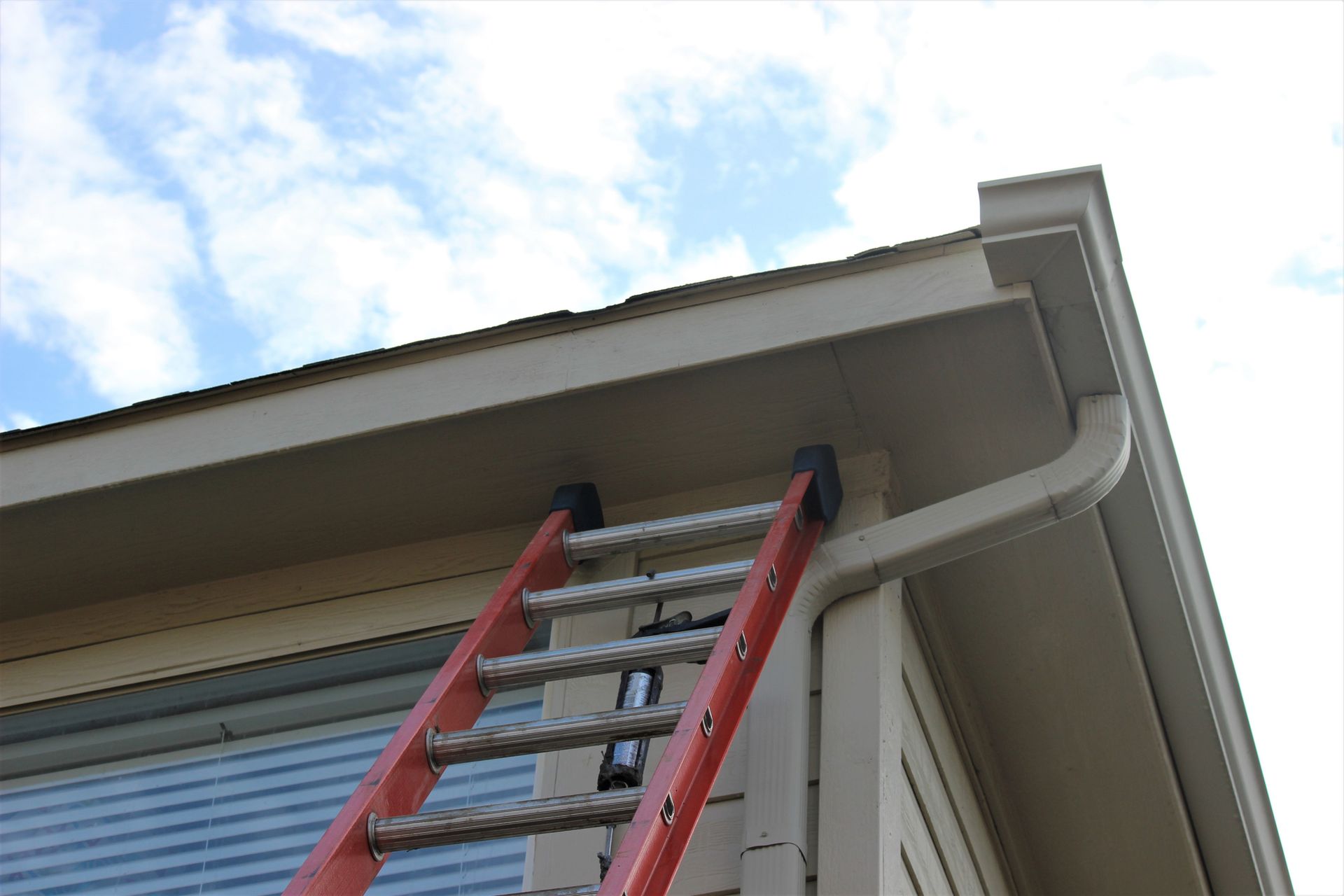 Red ladder against a tan house with a gutter, aiming towards the roof and cloudy sky.
