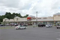 Strip mall storefronts with parked cars in a large parking lot under a cloudy sky