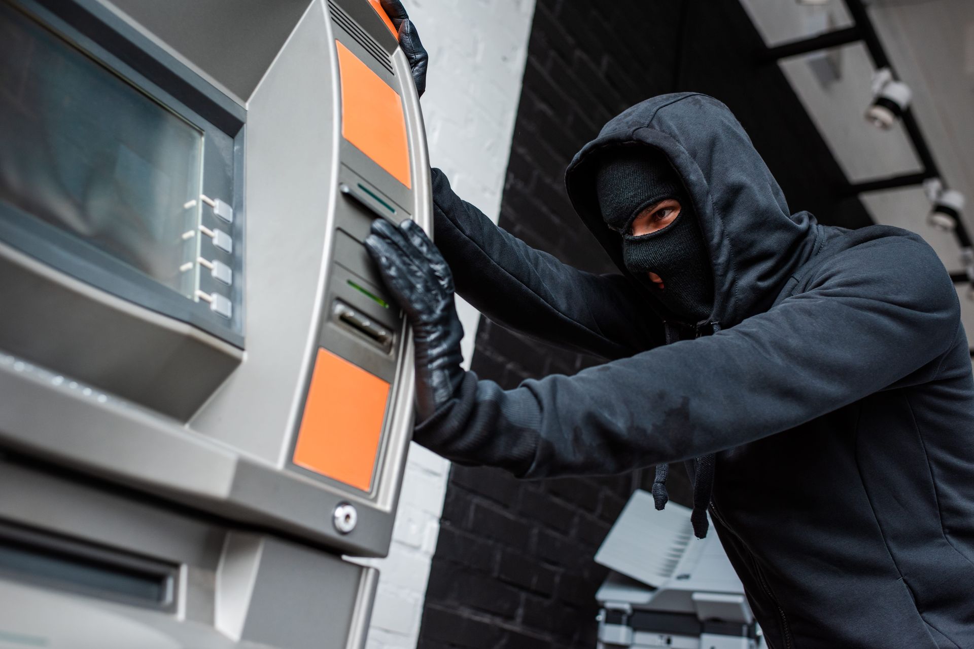 Masked burglar prying open an ATM with gloved hands, wearing a black hoodie.