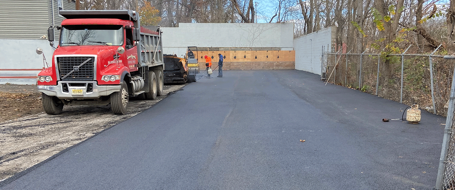 Red dump truck and workers paving a driveway beside a fence and trees