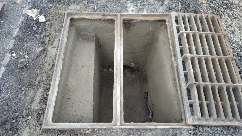Open concrete drainage pit with two rectangular compartments beside a metal grate on gravel.