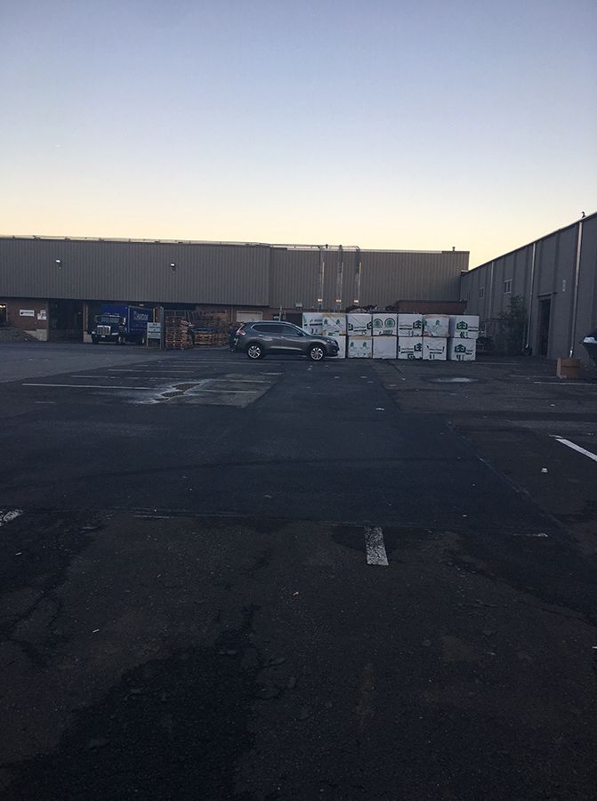 Empty parking lot beside industrial buildings at dusk, with a parked SUV and stacked crates.