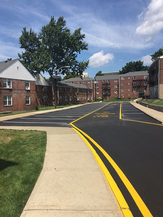 Campus road with fresh black asphalt, yellow lines, brick dorm buildings, and a partly cloudy sky.