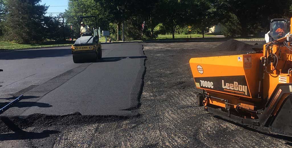 Road paving crew with orange machinery laying fresh black asphalt on a tree-lined street