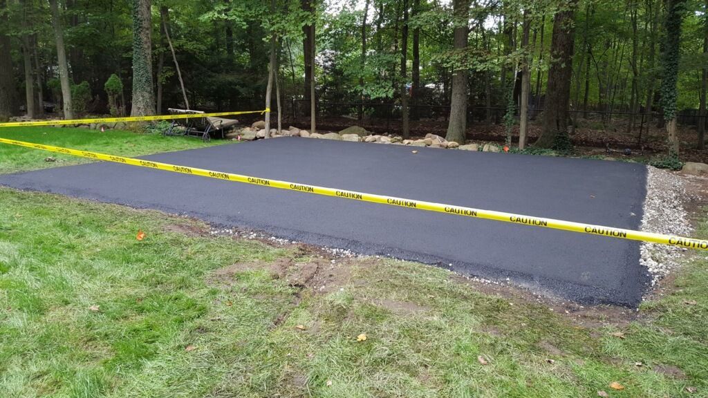 Freshly paved asphalt pad bordered by yellow tape in a wooded area.