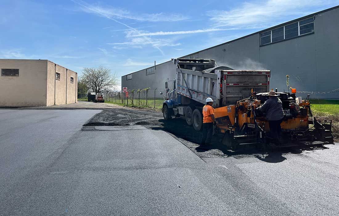 Snowplow spreading salt on an empty industrial parking lot beside a warehouse.