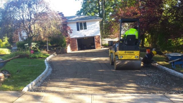 Yellow construction roller paving a sloped driveway toward a white house surrounded by trees and landscaping