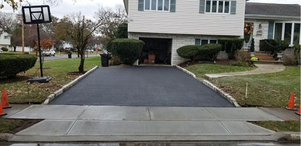 Driveway with a dark resurfaced path leading to a white house, bordered by wet concrete and orange cones