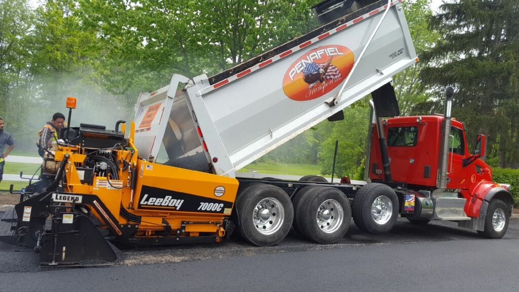 Orange paving machine dumps asphalt into red dump truck on a roadwork site