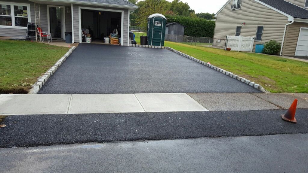 Freshly paved black driveway with a white concrete apron leading to a garage, orange cone at right, house in background