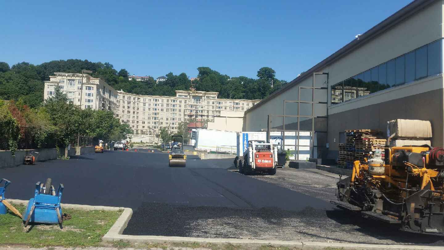 Road resurfacing at a building complex with workers, trucks, and paving equipment in the foreground