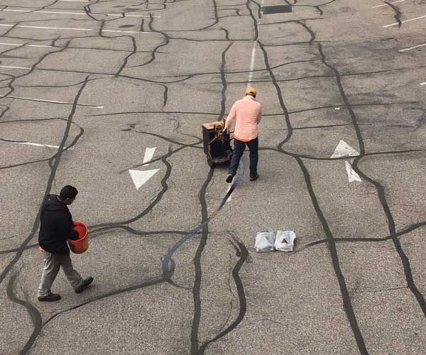 Two workers on a cracked asphalt road, one pushing a cart and one carrying a red bucket.