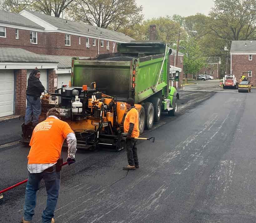 Road crew in orange safety gear operating a green street sweeper on a residential street