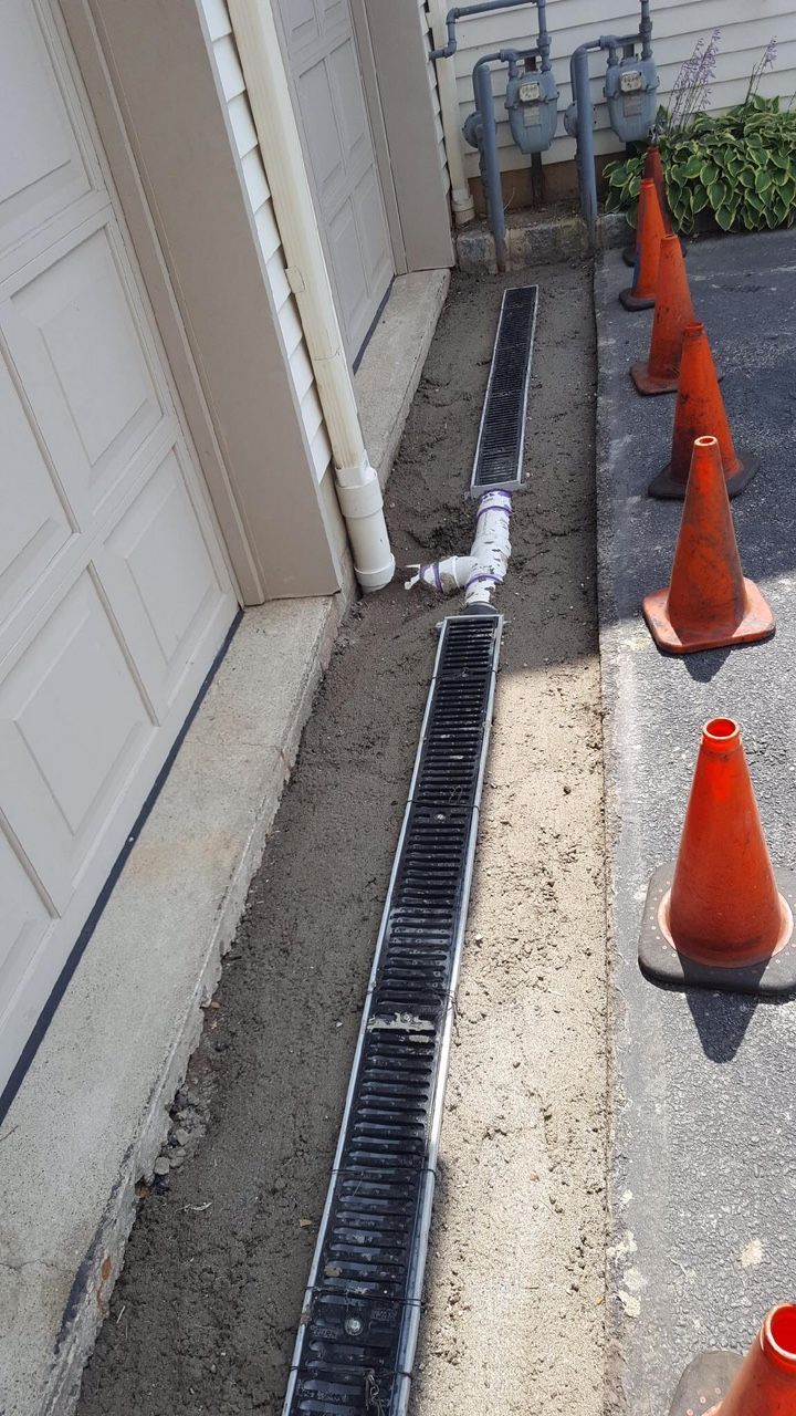 Narrow outdoor walkway beside a building, with drainage grates and orange traffic cones blocking the path