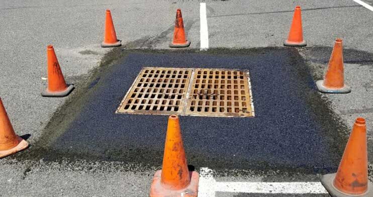 Orange cones surrounding a dark asphalt patch and metal storm drain grate in a road.