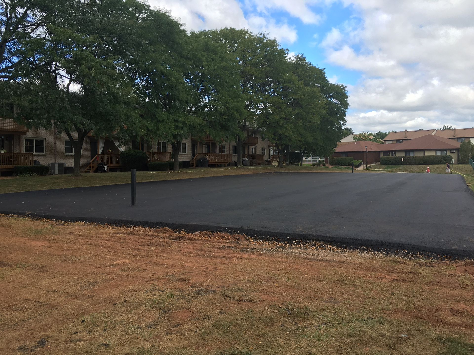 Empty asphalt lot beside a row of buildings and large trees under a partly cloudy sky