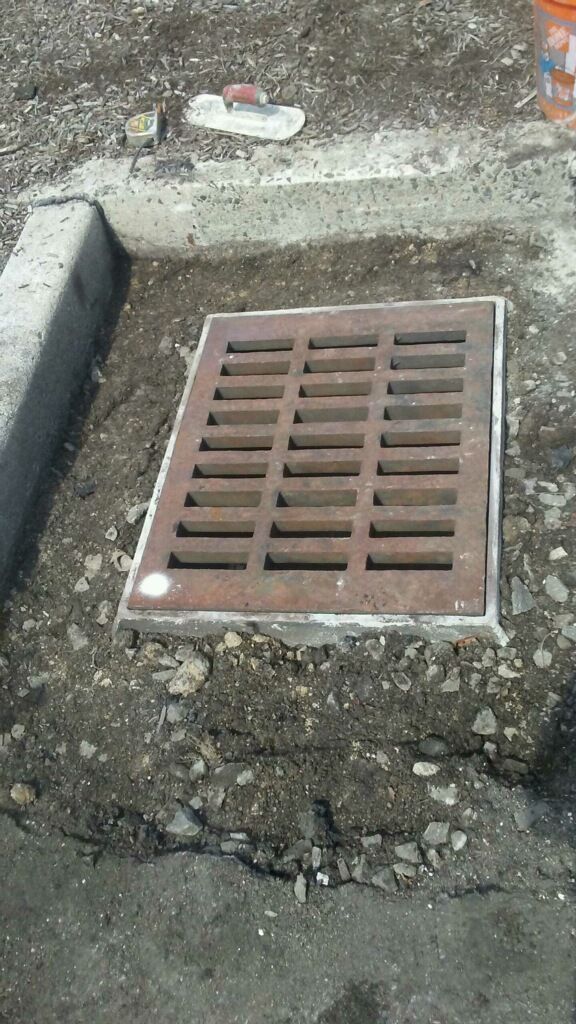 Rust-colored storm drain grate set in rough concrete and gravel ground.