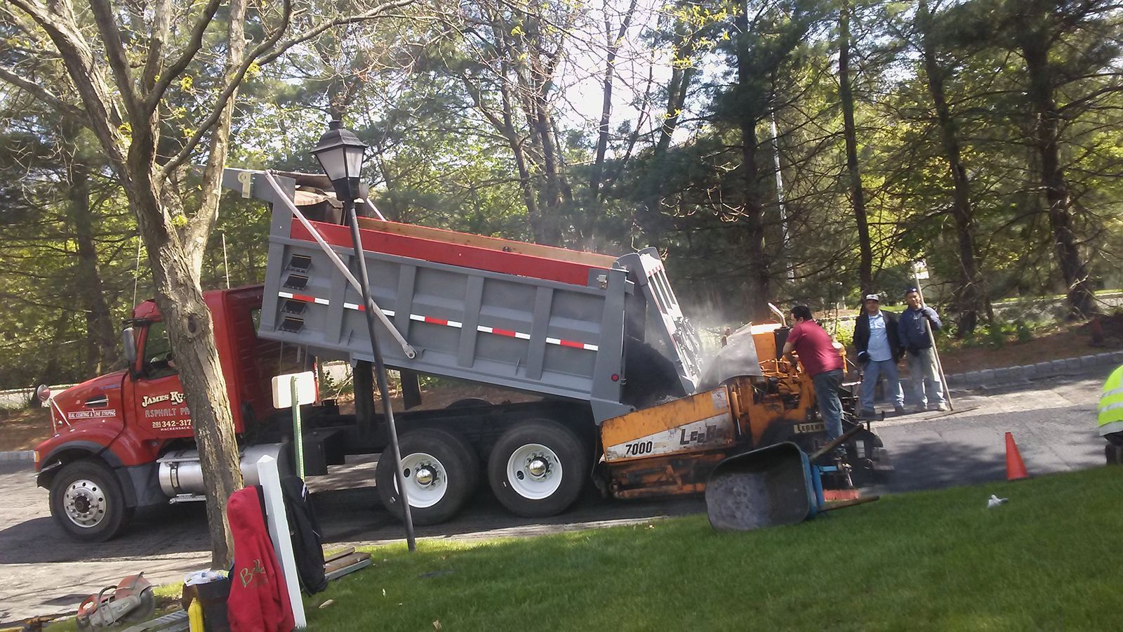 Workers and a red dump truck unloading asphalt at a roadside work site in a wooded area