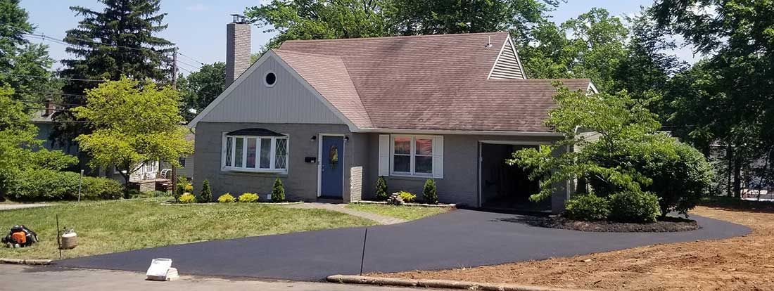 Small gray house with a brown roof, surrounded by trees and shrubs on a sunny day.