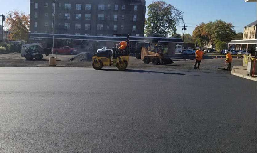 Construction workers paving a road beside parked vehicles and an apartment building in bright daylight